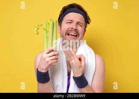 Lustiger sportlicher Mann mit Schnurrbart, der frischen Sellerie weinend hält. Ich hasse Gemüse, sollte aber für eine gesunde Ernährung essen. Studio an gelber Wand gedreht Stockfoto