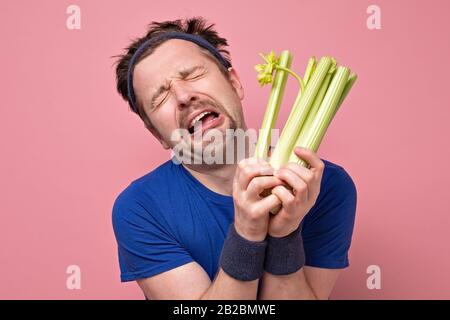 Lustiger sportlicher Mann mit Schnurrbart, der frischen Sellerie weinend hält. Ich hasse Gemüse, sollte aber für eine gesunde Ernährung essen. Studio an pinkfarbener Wand Stockfoto