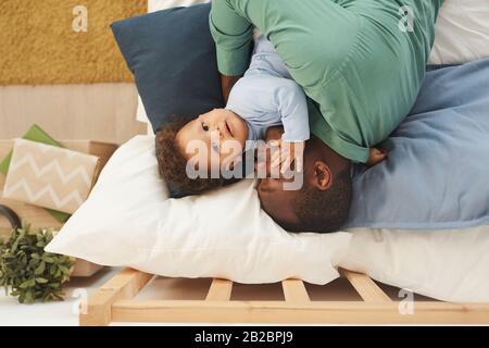 Oben sehen Sie das Porträt eines erwachsenen afro-amerikanischen Mannes, der Baby-Sohn umarmt, während er zu Hause auf dem Bett liegt, und kopieren Sie Platz Stockfoto