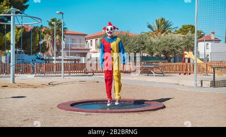 Ein gruseliger Clown, der ein farbenfrohes gelbes, rotes und blaues Kostüm trägt und auf einem Trampolin auf einem öffentlichen Spielplatz im Freien hüpft Stockfoto