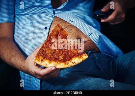 Nahaufnahme eines kaukasischen Mannes mit einem haarigen Bierbauch, auf dem Sofa sitzend, Pizza essen, während er den Fernseher beobachtet Stockfoto