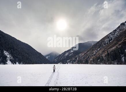 Einsame Frau läuft auf dem gefrorenen Kolsai-See mit Bergen im Hintergrund in Kasachstan Stockfoto