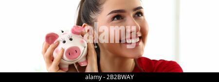 Portrait einer jungen, schönen Brunette-Frau, die eine pinkfarbene Ferkelbox gegen Geld hält Stockfoto