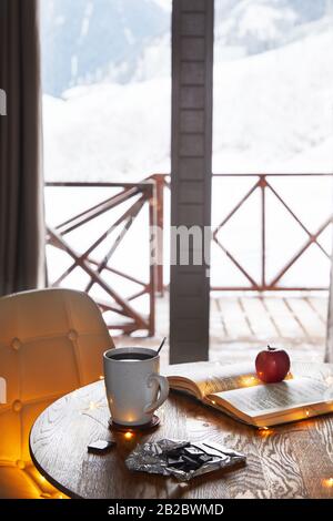 Kaffee, Schokolade, Buch und apfel auf dem Tisch in einem Winterhaus aus Holz mit wunderschönem Blick auf die Berge Stockfoto