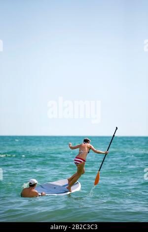 Frauen lernen, wie man ein SUP-Stehpaddelbrett verwendet, aber verlieren ihr Gleichgewicht und springen mit einem Paddel in der Hand ins Wasser. Tropischer heißer Tag Stockfoto