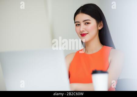 Erfolgreiche asiatische Geschäftsfrau trägt eine ärmellose orangefarbene Bluse, die mit einem Laptop und einer Kaffeetasse sitzt und Freude und Frische auf dem Schreibtisch weckt Stockfoto