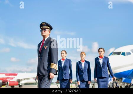 Portrait des reifen Piloten, der mit drei jungen, schönen Flugbegleitern gegen das Flugzeug auf dem Flughafen spazieren ging Stockfoto