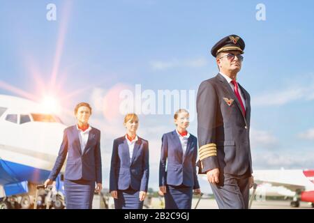 Portrait des reifen Piloten, der mit drei jungen, schönen Flugbegleitern gegen das Flugzeug auf dem Flughafen spazieren ging Stockfoto