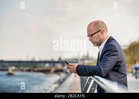 Porträt eines reifen Geschäftsmannes, der sich während der Verwendung des Smartphones an Geländer lehnt Stockfoto