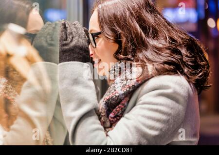 Porträt der jungen attraktiven Frau, die das Kleid auf einer Manequin im Laden durch das Fenster betrachtet Stockfoto