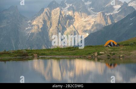 Touristen mit orangefarbenem Zelt in der Nähe des Bergsees. Felsen mit Schnee und Gletscher im Hintergrund. Stockfoto