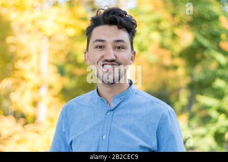 Gutaussehender junger Mann mit Blick auf die Kamera. Porträt des lachenden selbstbewussten und erfolgreichen jungen Mannes in einem blauen Hemd draußen. Fröhlicher Kerl lächelt Stockfoto