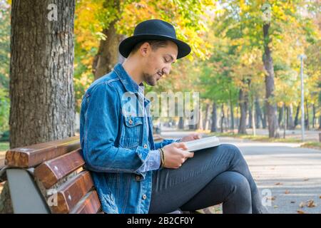 Gutaussehender junger Mann, der ein Buch in einem Park liest. Porträt eines jungen Mannes mit Denimjacke und blauem Hemd, das ein Buch draußen liest. Ein Kerl, der ein Buch i liest Stockfoto