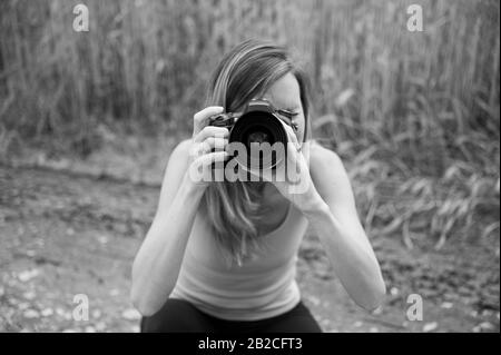 Monochromes Bild junger Fotografin, die in der Natur kniet und direkt an der Kamera fotografiert. Stockfoto
