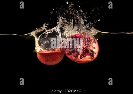 Köstliche Granatapfelhälften und spritzendes Wasser auf schwarzem Hintergrund Stockfoto