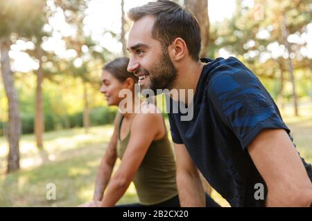 Foto eines netten lächelnden Paares in der Sportswear, das Übungen macht, während man im grünen Park während des sonnigen Sommers ausarbeitet Stockfoto