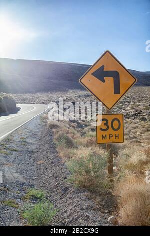 Gelbes Straßenschild zeigt eine Linksabbiegung an und gibt auf einer kurvenreichen Straße durch den Todes-Valley-Nationalpark eine Geschwindigkeitsbegrenzung von dreißig Meilen pro Stunde an Stockfoto