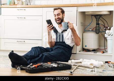 Bild des jungen positiven lächelnden Mannes Klempner arbeiten in einheitlichen Innenräumen mit dem Handy, das Kaffee trinkt. Stockfoto