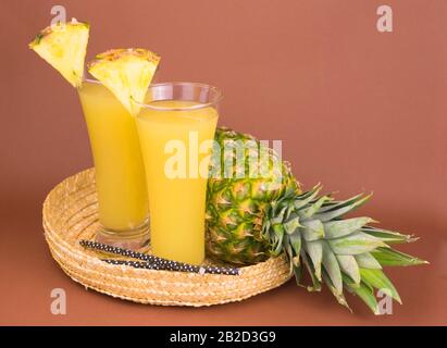 Zwei Gläser frischer Ananassaft und Ananas in einem Korbtablett auf dunkelrosa Hintergrund. Stockfoto