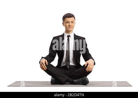 Junger Mann in einem schwarzen Anzug, der mit gekreuzten Beinen sitzt und auf einer Trainingsmatte meditiert, die auf weißem Hintergrund isoliert ist Stockfoto