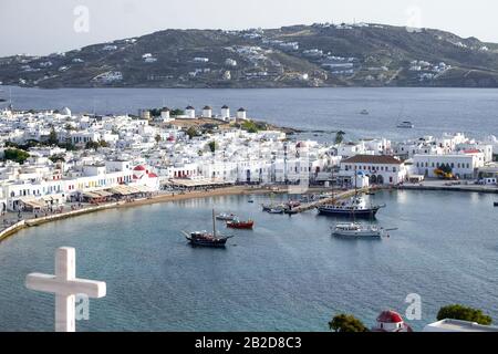 Blick auf den Sonnenuntergang über dem Stadthafen von Mykonos mit dem Kreuz der griechischen orthodoxen Kirche vor dem Hotel. Alte Windmühlen (Kato Mili) - berühmter Wahrzeichen und Touristenattraktion Stockfoto