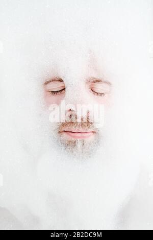 Männliches Gesicht in einem üppigen Schaum, nur Augen und Mund sind sichtbar, Nahaufnahme. Der Mann entspannt sich, während er in einer Badewanne liegt, mit seifigem Wasser. Draufsicht. Stockfoto