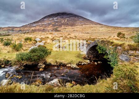 Alte kleine Steinbrücke über den Fluss mit Blick auf den Berg Errigal und stürmische Wolken, County Donegal, Irland Stockfoto