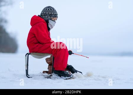 Kleiner Junge in roten Overalls beim Winterangeln. Sitzt auf einem Klappstuhl. In den Händen einer Angelrute. Blickt in die Ferne. Weißer Schnee. Die Skyline verschmilzt mit dem Himmel. Stockfoto