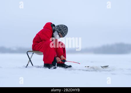 Kleiner Junge in roten Overalls am See. Winterangeln. Angelrute in den Händen eines Jungen. Er hat sich nach unten gebogen und schaut vorsichtig in das Loch. Weißer Schnee rundum. Niedrige Wolken. Überwiegend bewölkt. Stockfoto