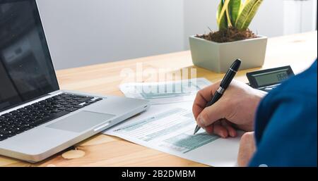 Über der Schulter Nahaufnahme des Mannes, der das US-Steuerformular 1040 von Hand und Stift mit Rechner und Laptop im Büro berechnet und füllt. Stockfoto