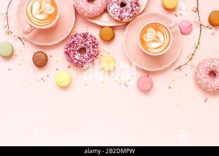 Zwei Latte Coffee Cups, köstliche pinkfarbene Doughnuts mit Einstreuung und bunte helle Makronen auf pinkfarbenem Papierhintergrund Stockfoto
