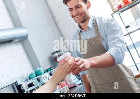 Kaffeepassion. Der junge Barista im Coffeeshop serviert dem Kunden eine Tasse heißen Kaffee und lacht fröhliche Nahaufnahme Stockfoto
