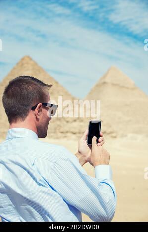 Der Geschäftsmann, der sein Smartphone in der Wüstensonne vor dem Hintergrund der Großen Pyramiden in Ägypten benutzt Stockfoto