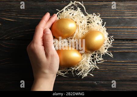 Weibliche Hand hält Gelege mit goldenen Eiern auf Holzhintergrund, Draufsicht Stockfoto