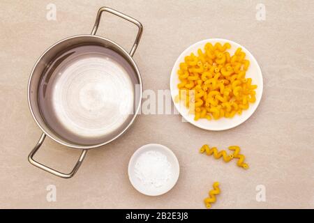 Roher Fuzilli mit Wasser in Topf und Salz. Traditionelle italienische Pasta in weißer Platte auf Steingrund. Vorbereitung für Kinder, Draufsicht Stockfoto