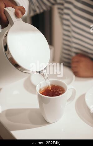 Teeparty in einer hellen Küche mit weißem Tisch und Stühlen. Schokoladenkuchen mit Mandeln im Vordergrund, neben einer weißen Tasse mit heißem Tee und Silve Stockfoto