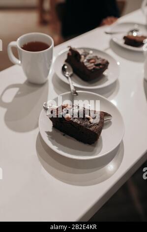 Stück Schokoladenkuchen mit Mandeln in einer hellen Küche, Tisch mit weißem Becher mit heißem Tee und silberfarbenem Besteck Stockfoto