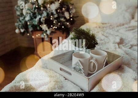 Zwei weiße Tassen auf einem Holztablett stehen auf dem Bett mit einer flauschigen Decke vor dem Hintergrund der Weihnachten, bokeh aus Girlanden im Vordergrund Stockfoto