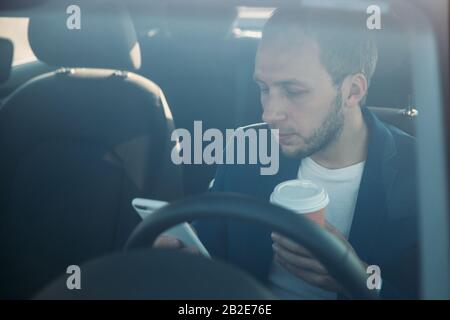 Vorderansicht des jungen Geschäftsmannes, der im Auto sitzt und auf dem Smartphone tippt/nachgibt, Kaffee in einer Papierbecher trinkt, durch die Windschutzscheibe gesehen. Kaffee B. Stockfoto