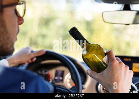 Nahaufnahme des betrunkenen Mannes, der mit einer Flasche Alkohol in der Hand ein Auto fährt und dabei das Lenkrad hält.Trunkenheit. Trinken Sie nicht zum Autofahren. Stockfoto