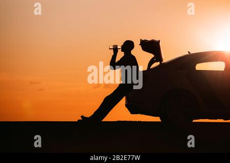 Silhouette des Mannfahrers nach einer Fahrt entspannend, auf dem Kofferraum seines Autos sitzend und Trinkwasser aus einer Flasche, Seitenansicht. Sunset Time. Stockfoto