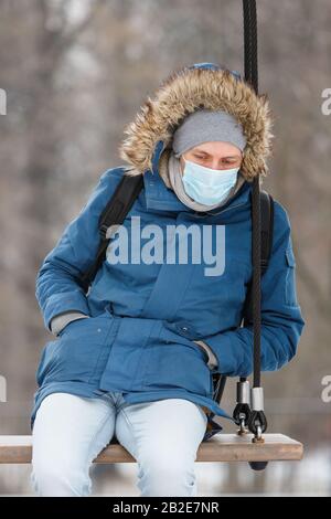 Kranker Mann mit einer Haube, die allein auf einem Schwingen sitzt, eine Gesichtsmaske gegen übertragbare Infektionskrankheiten trägt und gegen den schützt Stockfoto