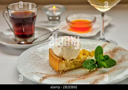 Stück Kuchen charlotte mit Äpfel-Eisball und frischer Minze auf weißem Teller. Serviert mit einer Rosette Honig, einer Tasse aromatischen Tees und einem Glas Stockfoto