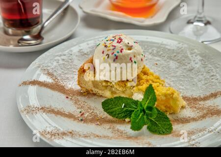Stück Kuchen charlotte mit Äpfel-Eisball und frischer Minze auf weißem Teller. Serviert mit einer Rosette Honig, einer Tasse aromatischen Tees und einem Glas Stockfoto