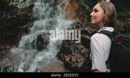Mädchen Backpacker Blick auf Dschungel Cascade Wasserfall. Fröhliche Kaukasische Frau, Aktive Reisende Discovery Wilde Thailand Provinz Natur. Tourist Contemplating Tropical River Flow, Smile. Stockfoto