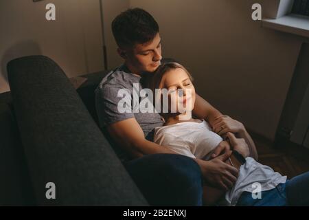 Charmantes blondes Mädchen, das mit geschlossenen Augen auf ihrem kaukasischen Liebhaber liegt, während sie zusammen auf einem Sofa sitzen Stockfoto