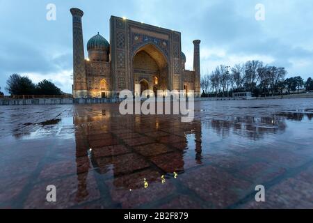 Sher Dor Madrasa mit Spiegelung in Pfütze an einem kalten Winterregnertag, Registan, Samarkand, Usbekistan Stockfoto