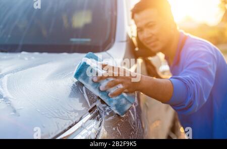 Asiatischer Mann mit blauem Schwamm und Seife zum Waschen des Autos im Freien bei Sonnenuntergang. Konzept für die Reinigung und Wartung von Autos Stockfoto