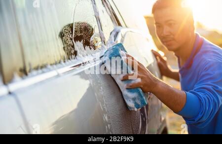 Asiatischer Mann mit blauem Schwamm und Seife zum Waschen des Autos im Freien bei Sonnenuntergang. Konzept für die Reinigung und Wartung von Autos Stockfoto