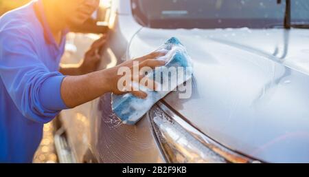 Asiatischer Mann mit blauem Schwamm und Seife zum Waschen des Autos im Freien bei Sonnenuntergang. Konzept für die Reinigung und Wartung von Autos Stockfoto